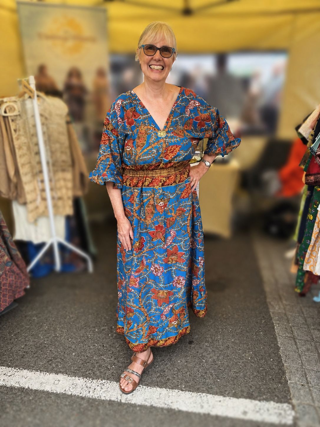 A woman wearing a blue bohemian floral maxi dress with a matching waist belt, standing in what appears to be a market or boutique setting.