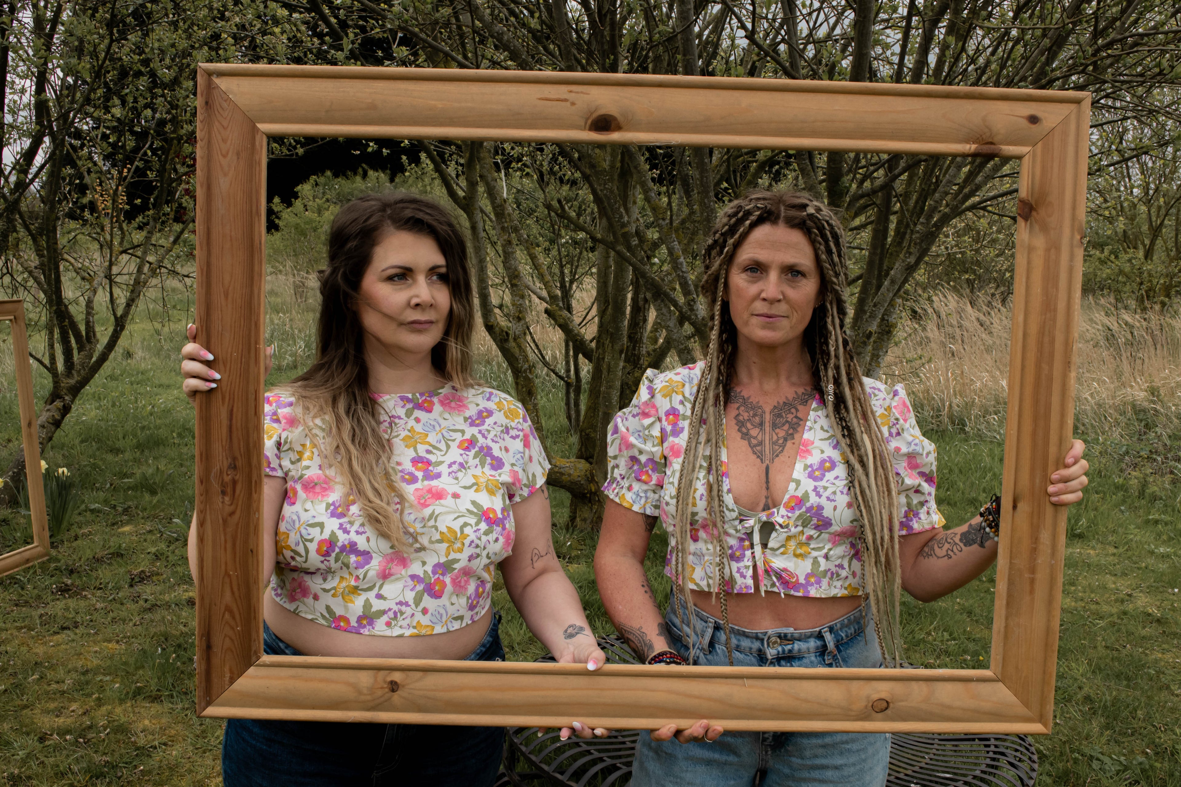Two women holding a wooden picture frame outdoors with trees in the background