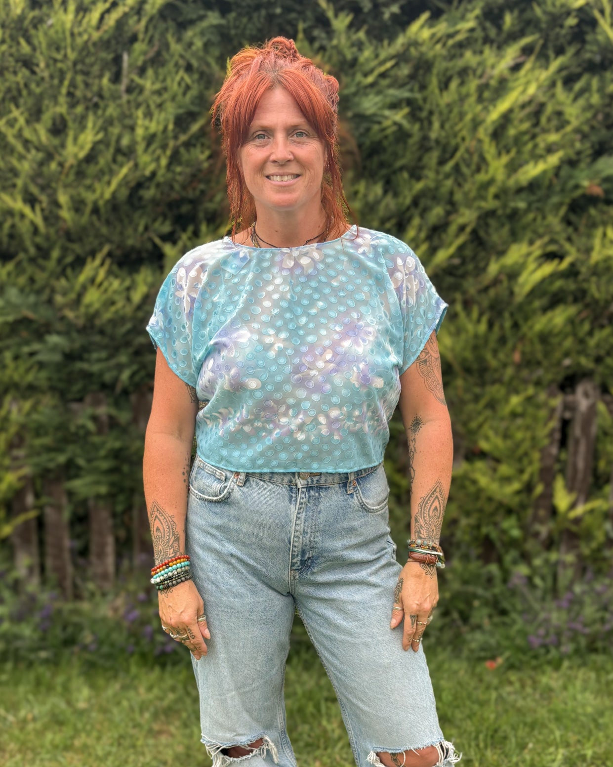 Person wearing a light blue patterned top and ripped jeans standing in a grassy area with greenery.