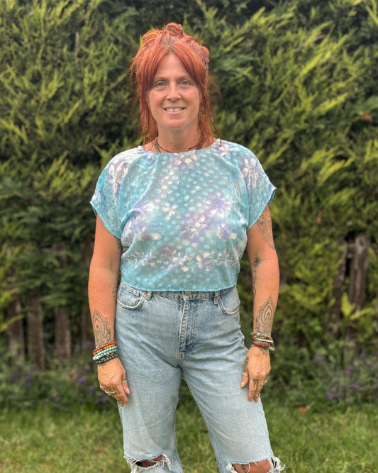 Person wearing a light blue patterned top and ripped jeans standing in a grassy area with greenery.