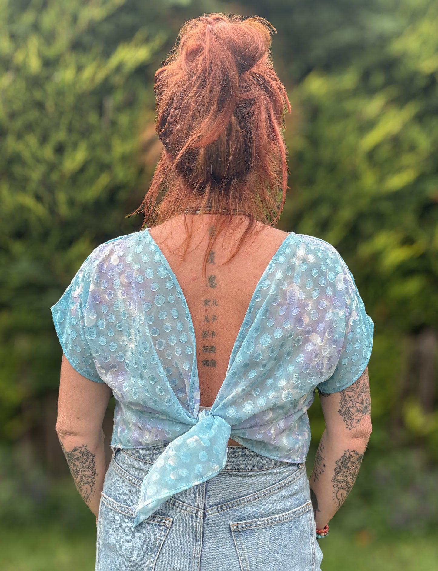 Person wearing a light blue patterned top with a tied front, standing outdoors with greenery in the background.