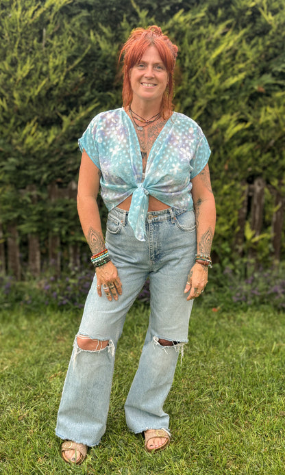Person with red hair wearing a light blue top and ripped jeans standing in a grassy area with greenery.