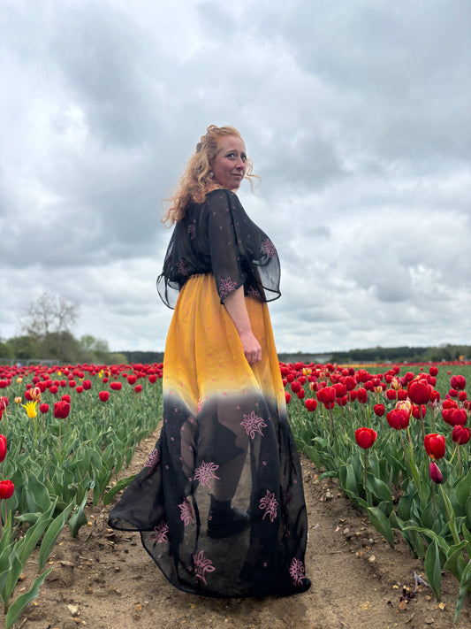 A woman standing in a field of red tulips at tulips for tapping wearing a black and gold long dress with a deep V-neckline and front split.