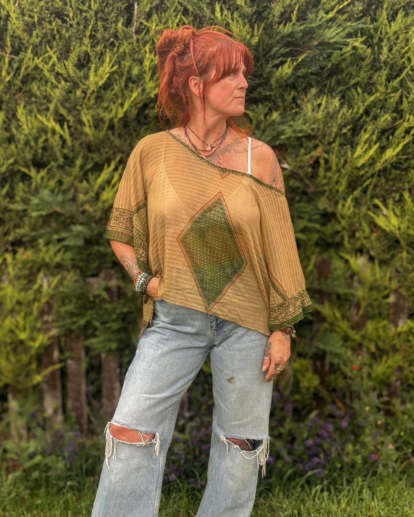 Woman wearing a mustard yellow top with a green geometric pattern and ripped jeans standing in a garden.