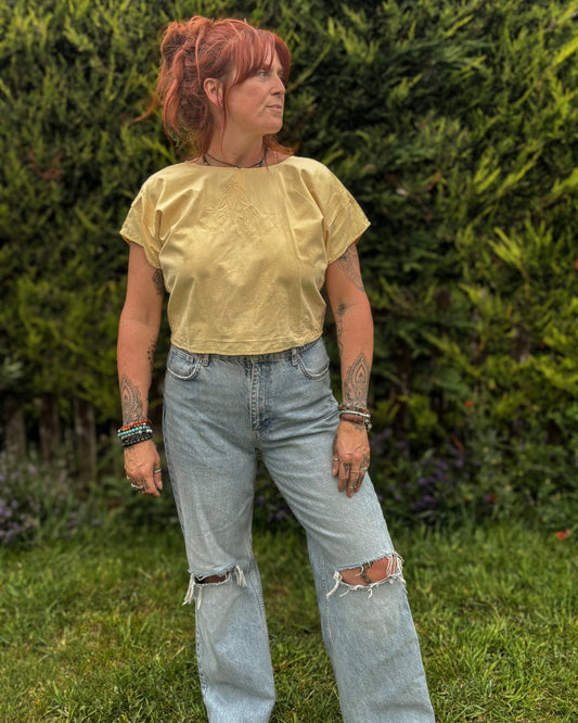 Person wearing a yellow top and blue jeans standing outdoors with greenery in the background