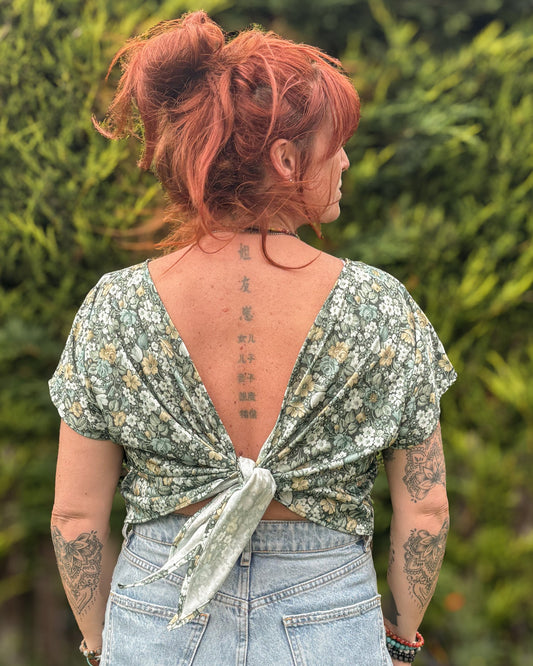 Person with red hair tied back, wearing a floral top and denim shorts, standing against a green bush background.