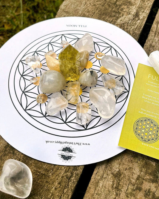 A crystal grid with various crystals arranged on it, placed on a paper with a floral design, next to a yellow ritual card and a small clear crystal.