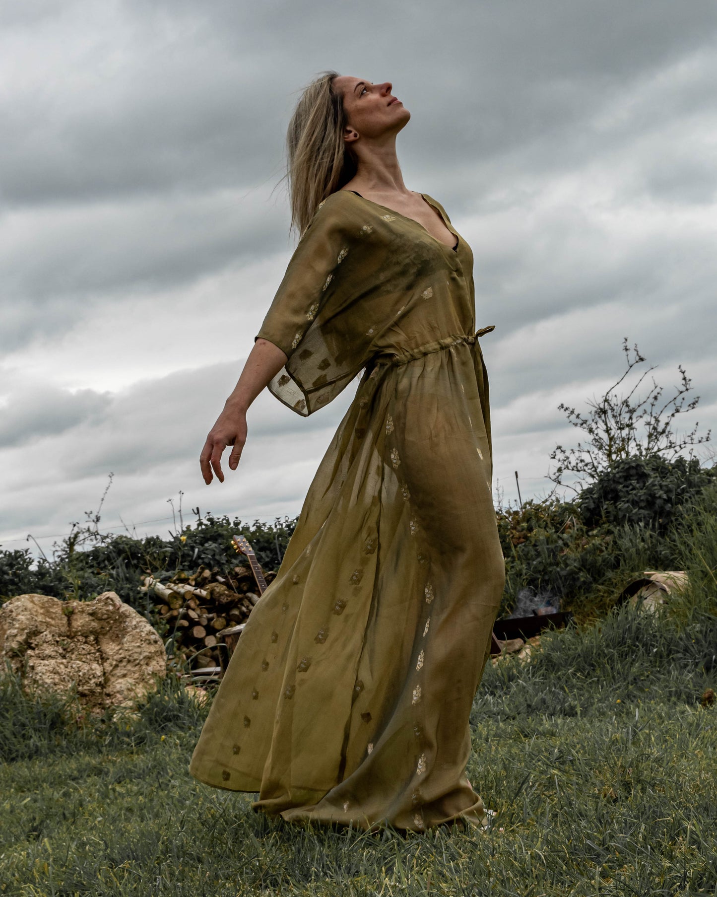 A person standing in an outdoor setting wearing a long, green, sleeveless dress with a deep V-neck and front split.