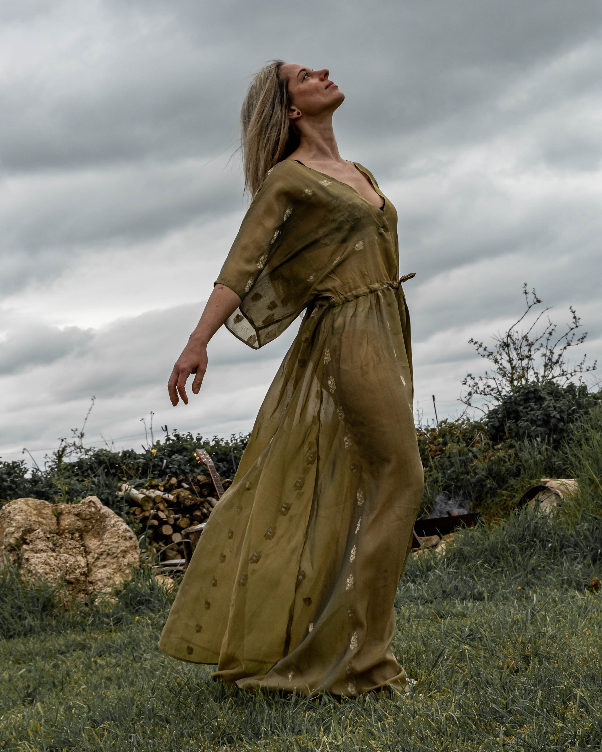 A person standing in an outdoor setting wearing a long, green, sleeveless dress with a deep V-neck and front split.
