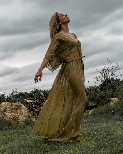 A person standing in an outdoor setting wearing a long, green, sleeveless dress with a deep V-neck and front split.