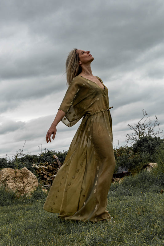 A person standing in an outdoor setting wearing a long, green, sleeveless dress with a deep V-neck and front split.