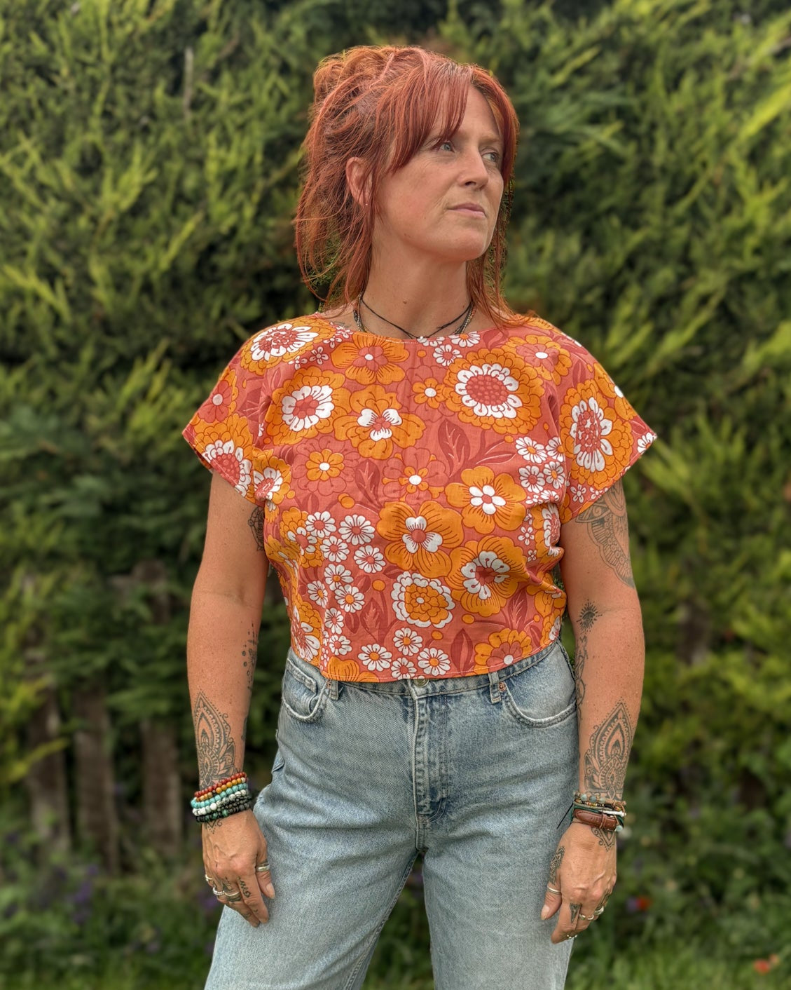Person wearing a floral top and ripped jeans standing outdoors with greenery in the background