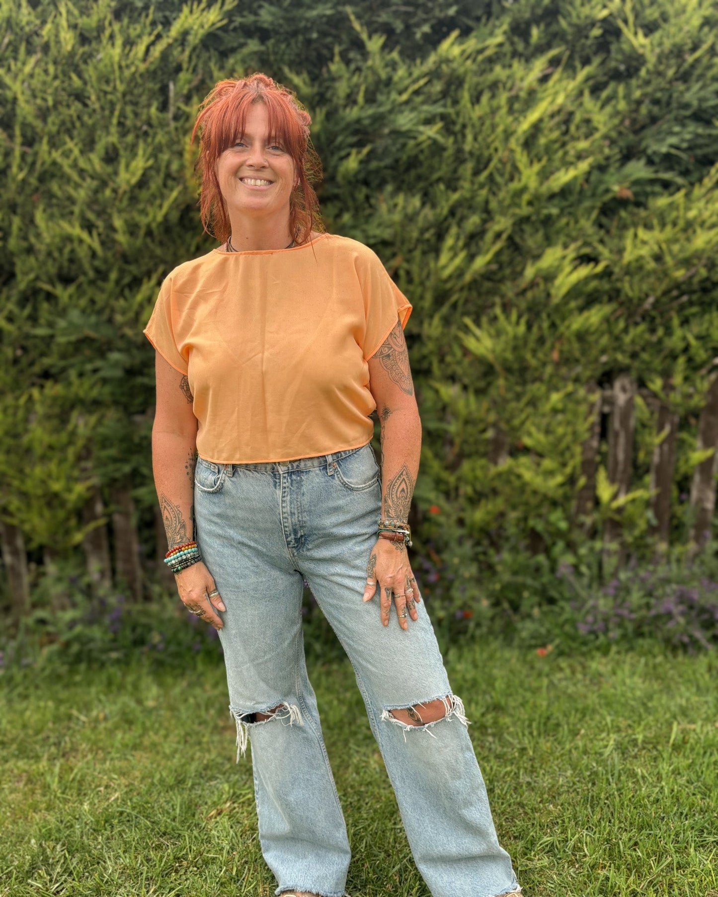 Woman wearing an orange top and light blue jeans standing in a grassy area with greenery.