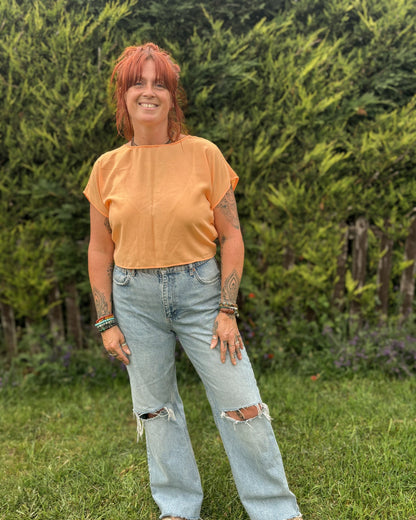 Woman wearing an orange top and light blue jeans standing in a grassy area with greenery.