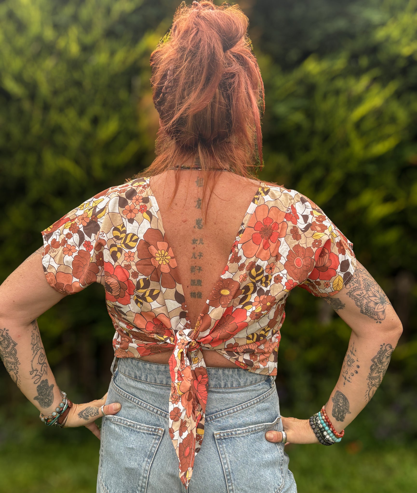 Person wearing a floral top with an open back, standing outdoors with greenery in the background.
