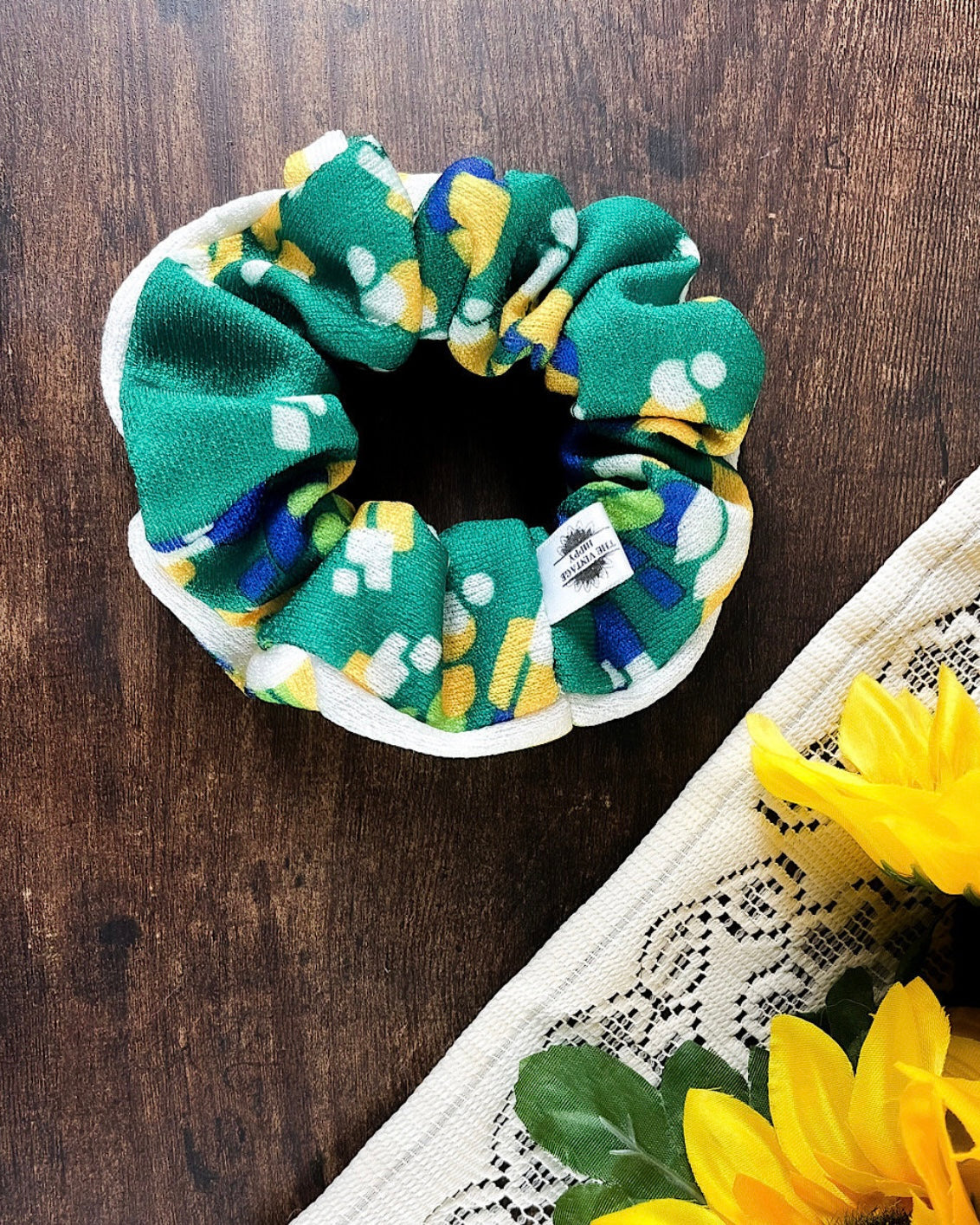 A green, blue, and yellow dotted scrunchie on a white band, placed on a wooden surface with a sunflower in the background.