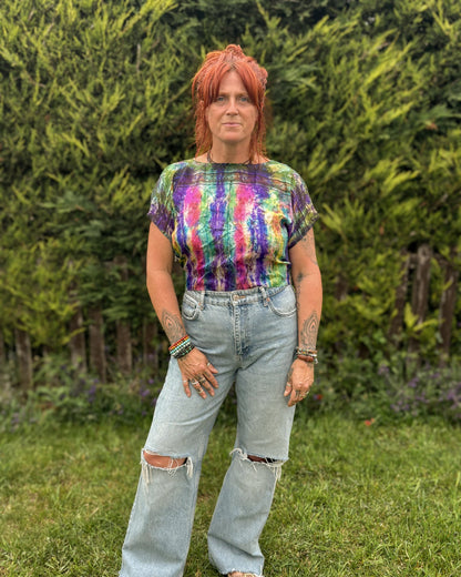 Person wearing a tie-dye shirt and ripped jeans standing outdoors with greenery in the background