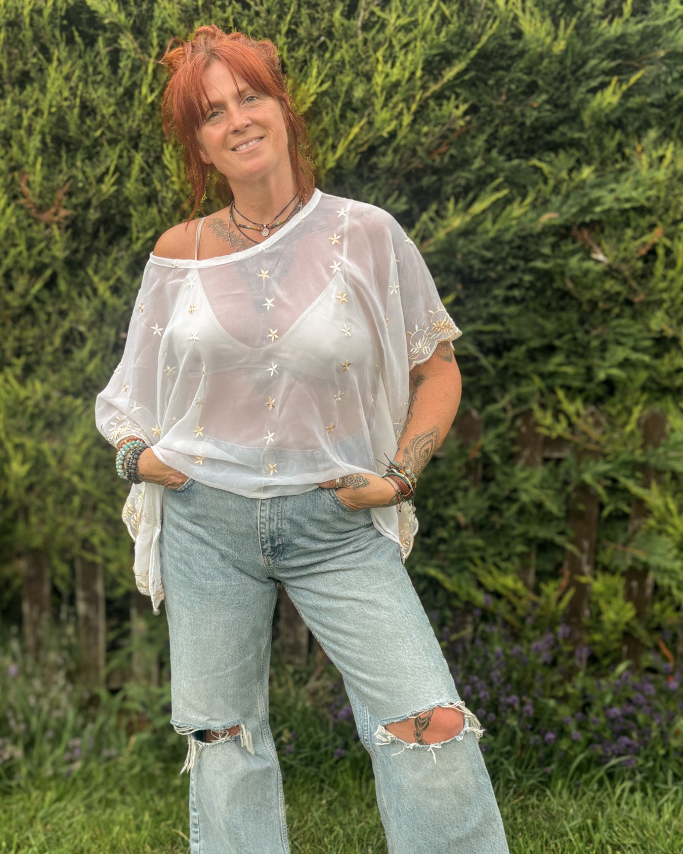 Person wearing a sheer top and ripped jeans standing outdoors with greenery in the background