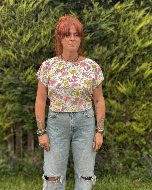 Person wearing a floral top and ripped jeans standing outdoors with greenery in the background
